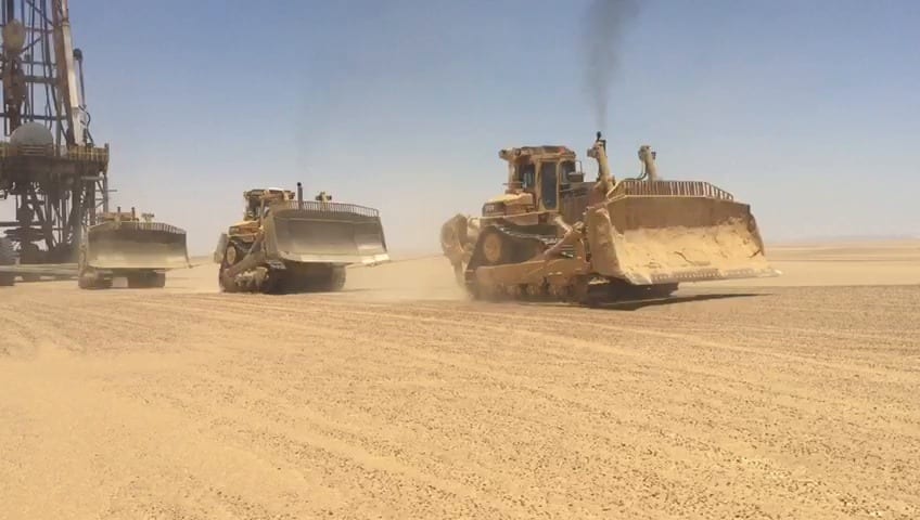Bulldozers towing a drilling rig across the desert.